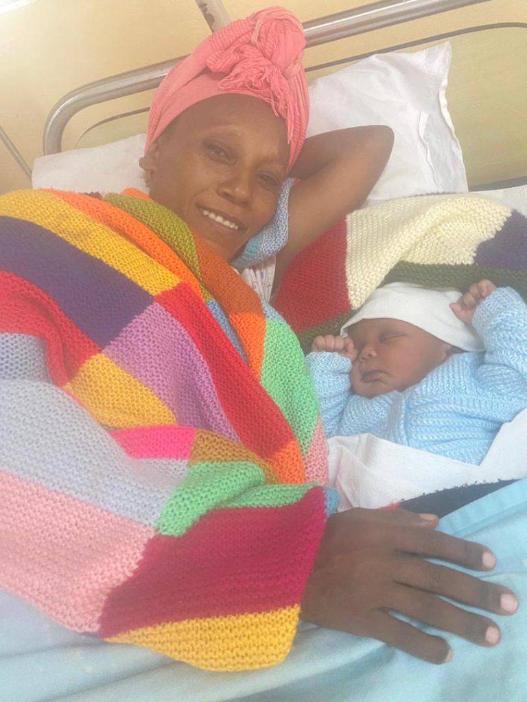 A mother wrapped in a colourful blanket lies beside her newborn baby